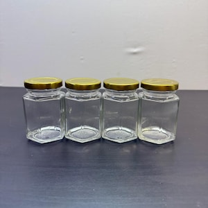 Peut inclure: Quatre bocaux en verre transparent avec des bases hexagonales et des couvercles dorés sont disposés en rangée. Les bocaux sont vides et semblent neufs. Les bocaux sont adaptés au stockage d'aliments.