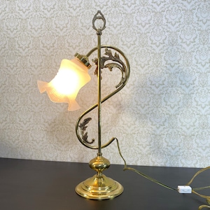 May include: A vintage brass table lamp with a frosted glass shade. The lamp has an ornate, curved design with floral accents. The base and stem are made of polished brass. The shade is a soft, off-white colour, and the lamp is lit.