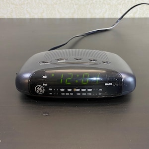 May include: A black GE digital alarm clock showing 12:04 AM in green digits. The clock has buttons for setting time and radio. It also displays radio frequencies in MHz.