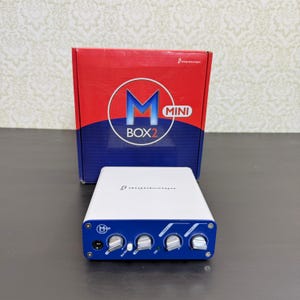 May include: A white and blue Digidesign Mbox 2 Mini audio interface with four silver control knobs on the front panel, positioned in front of its original red and blue cardboard packaging box featuring the Mbox 2 Mini logo and text.