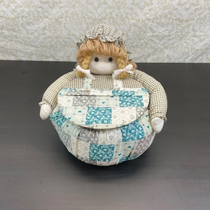 May include: A fabric doll featuring a round body with a patchwork apron in shades of teal, white, and grey. The doll has blonde yarn hair styled in braids, a matching gingham bonnet, and white ribbons tied at the base of each braid on the head.