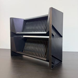 May include: A black, geometric, two-tiered storage unit. Each tier features a series of vertical, wavy dividers. The unit has a modern, angular design with a dark finish, suitable for organizing items.