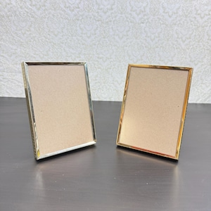 May include: Two rectangular picture frames with thin, polished gold-tone metal borders. Each frame features a simple, clean-lined design with a neutral-colored backing board visible inside, standing upright to display photographs or artwork.