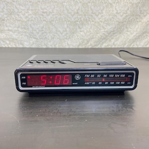 May include: A black GE digital clock radio featuring a red LED display showing 5:06, an AM/PM indicator, and a manual analogue radio tuner dial for FM and AM frequencies. The device includes a top-mounted speaker grille and control buttons.