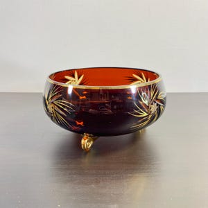 Puede incluir: Un cuenco de vidrio marrón oscuro con detalles dorados y un diseño geométrico. El cuenco tiene tres patas doradas.