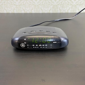 May include: A black GE digital alarm clock displaying 12:00 in green numerals. The clock has a rectangular shape with rounded edges, a speaker grill on top, and a series of buttons. The clock is plugged in with a black power cord.