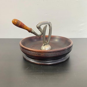 May include: A vintage nutcracker with a wooden handle and a metal cracking mechanism, set in a dark brown wooden bowl. The bowl has a tiered design. A classic kitchen tool for cracking nuts.