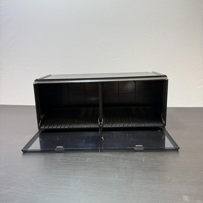 May include: A black, two-compartment bread box with clear, hinged doors. The box is rectangular with a flat top and sits on a dark surface. Each compartment has a series of parallel ridges on the bottom.