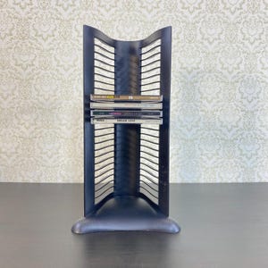 May include: A black metal CD rack with a curved top and a slotted design. Several CDs are visible in the rack, with titles including "Dream Love" and "Madonna". The rack is on a dark surface against a patterned wallpaper.