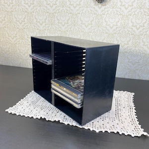 May include: A black CD storage unit with two compartments, each with slots for organising discs. Several CDs are visible inside. The unit sits on a white, crocheted doily, with a patterned wallpaper background.