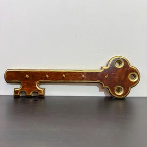 May include: A wooden key-shaped key holder for wall mounting. The key is dark brown with gold-coloured trim and key hooks. The key holder has a classic design and is suitable for home decor.