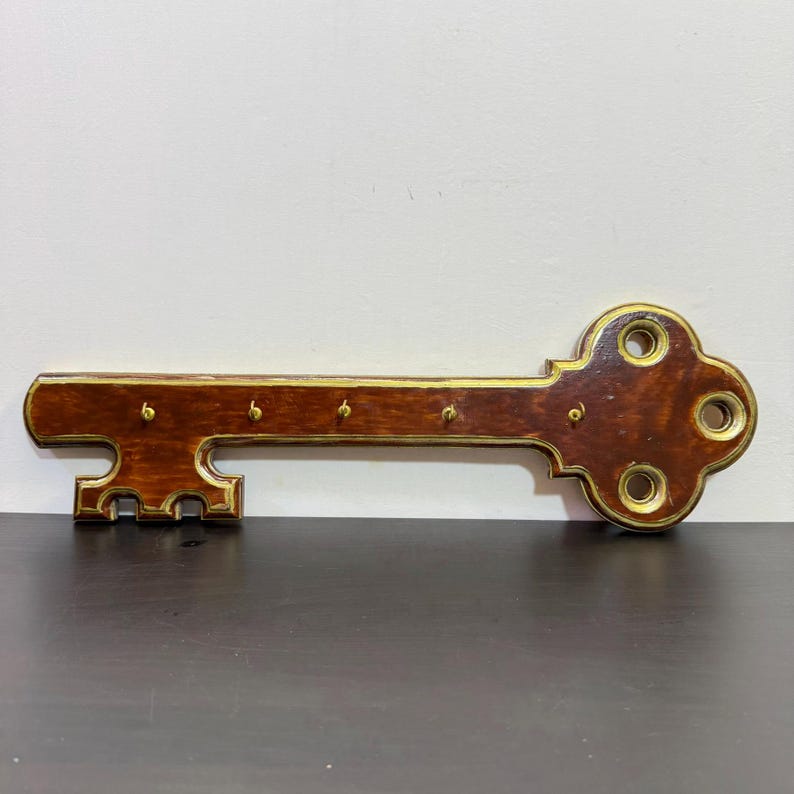 May include: A wooden key-shaped key holder for wall mounting. The key is dark brown with gold-coloured trim and key hooks. The key holder has a classic design and is suitable for home decor.