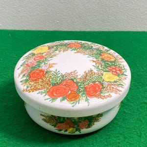 May include: White ceramic lidded trinket box with a floral design. The lid features a wreath of red, yellow, and orange flowers with green leaves. The base of the box has a similar floral design.