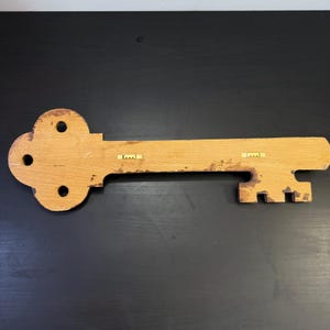 May include: A large, wooden key-shaped wall decoration. The key is made of light-coloured wood with a distressed finish. It features a decorative head with three circular cutouts and a long, rectangular shaft. Two metal hangers are attached to the back for mounting.