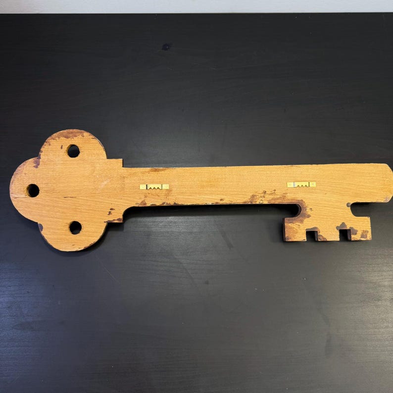 May include: A large, wooden key-shaped wall decoration. The key is made of light-coloured wood with a distressed finish. It features a decorative head with three circular cutouts and a long, rectangular shaft. Two metal hangers are attached to the back for mounting.