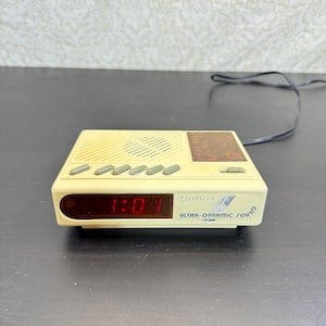 May include: A cream-colored Citizen alarm clock radio with a digital display showing 1:01 in red. The device features a top-mounted speaker grille, five grey control buttons, a radio tuning dial, and a black power cord extending from the back.