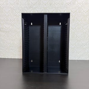 May include: A black metal media storage rack featuring two vertical columns with multiple horizontal slots for organizing discs. The unit has a rectangular frame with four keyhole-shaped mounting holes on the back panel for wall installation.