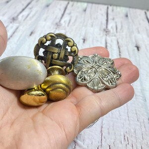 Op de afbeelding: Een verzameling van vier decoratieve kastknoppen. Twee zijn goudkleurig, één is zilverkleurig en één is een wit ovaal. De knoppen hebben ingewikkelde ontwerpen.