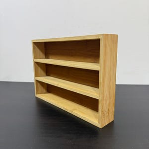 May include: A small, three-tiered bamboo shelf. The natural wood-toned shelf has three horizontal shelves and is designed to sit on a flat surface. The shelf is made of bamboo and has a simple, minimalist design.