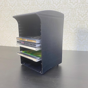 May include: A black CD storage unit with multiple shelves, holding several compact discs. The unit has a curved top and a rectangular base. The CDs are visible on the shelves, with their cases and labels showing. The unit is made of a solid, dark material.