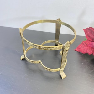 Może przedstawiać: Zabytkowy, mosiężny, dekoracyjny stojak z okrągłym blatem i ozdobnymi detalami kwiatowymi. Stojak ma trzy nogi i jest przeznaczony do trzymania okrągłego przedmiotu. Mosiądz ma ciepły, złoty odcień.