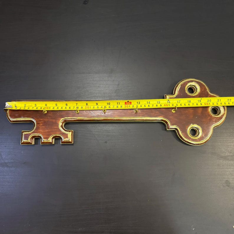May include: A decorative key-shaped wall-mounted key holder. The key is made of brown wood with gold trim and has several hooks for keys. The key measures approximately 56 cm in length.