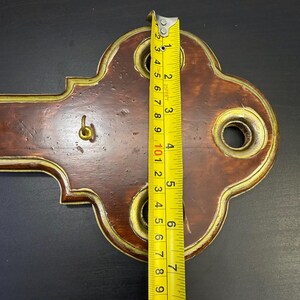 May include: A decorative wooden wall key holder with brass accents. The key holder is approximately 23 cm long. It features a hook for keys and three decorative holes. The wood has a dark brown finish with a gold trim.