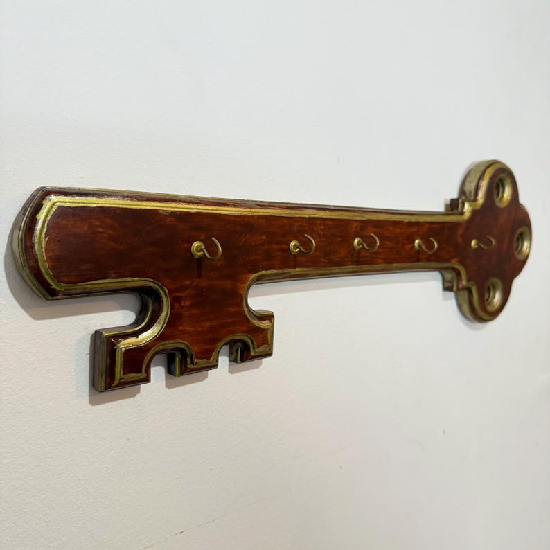 May include: A wooden key holder in the shape of a large key, mounted on a wall. The key is dark brown with gold edging and has five hooks for keys. The wall is painted white.