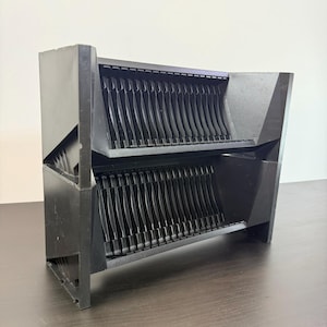 May include: A black, two-tiered dish rack with a geometric design. The rack features vertical slots for plates and bowls, and is made of a matte black material. The rack is designed to sit on a countertop.