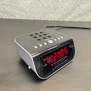 May include: A black and silver digital alarm clock radio with a red LED display showing 12:05. The top surface features a grid of circular speaker holes and multiple control buttons labeled for time, alarm, and radio functions with a power cord.