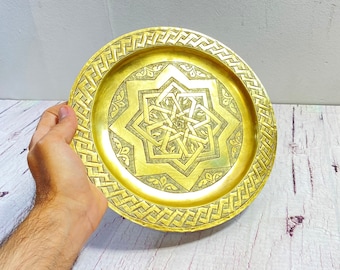 May include: A round, gold-colored metal plate with an intricate geometric star pattern in the center. The plate has a decorative border with a woven design. The plate is held by a hand.