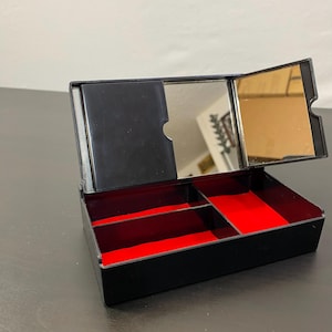 May include: A black jewellery box with a red velvet interior. The box has a hinged lid with two mirrors and several compartments for storing jewellery. The box is open, revealing the interior.