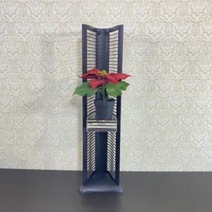 May include: A dark gray CD rack with a potted poinsettia plant and a stack of CDs. The rack has a vertical, open design with multiple slots for CDs. The poinsettia has red bracts and green leaves.