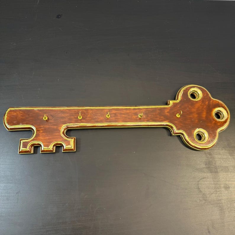 May include: A decorative key-shaped wall-mounted key holder. The key holder is made of dark brown wood with gold-colored trim. It features five small hooks for hanging keys and decorative holes. The key measures approximately 38 cm in length.