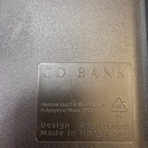 May include: A close-up of a gray plastic CD case with the text "CD BANK" embossed on the surface. The case also includes text indicating the material used is polystyrene plastic and that it was made in Hong Kong.