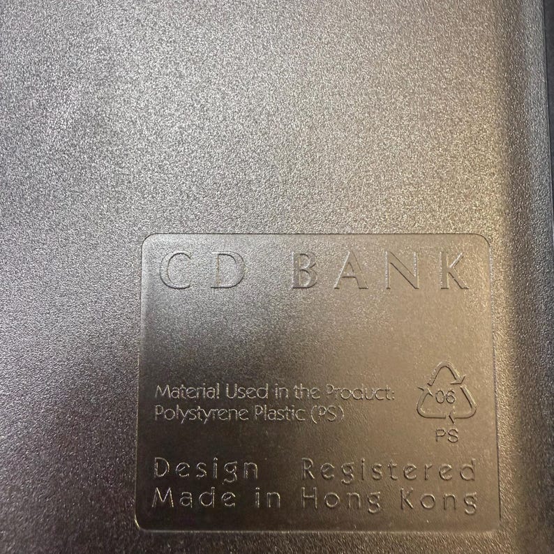 May include: A close-up of a gray plastic CD case with the text "CD BANK" embossed on the surface. The case also includes text indicating the material used is polystyrene plastic and that it was made in Hong Kong.