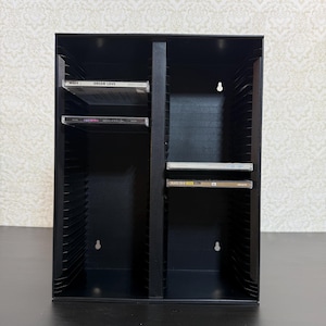 May include: A black metal CD storage rack featuring two vertical columns with multiple horizontal slots for organizing discs. The unit includes keyhole-shaped mounting holes on the back panel for wall installation and contains four CD cases.