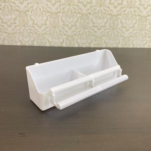 May include: A white plastic bird feeder with a rectangular shape and two compartments. It features a perch and is designed to attach to a cage. The feeder is clean and simple, suitable for small birds.
