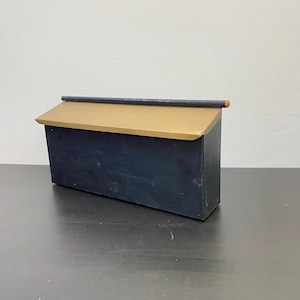 May include: A dark blue wooden box with a gold-coloured slanted roof. A dark rod with a small orange end piece sits on top of the roof. The box is rectangular and sits on a dark surface.
