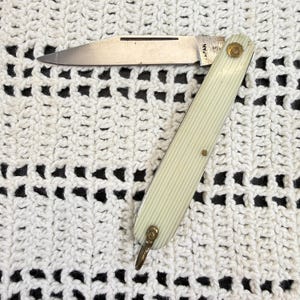 Puede incluir: Una navaja plegable con una hoja de metal plateado y un mango de color crema con una textura acanalada vertical. La hoja tiene grabado el texto JAPAN. En la base del mango hay una pequeña anilla de metal de color latón.