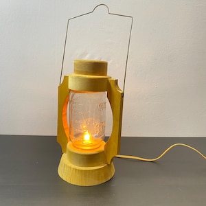 Pode incluir: Uma lanterna de madeira amarela com um frasco de vidro e uma luz dentro. A lanterna tem uma alça de metal e um cabo.