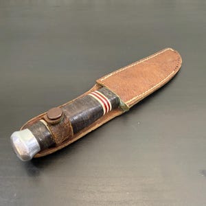 Peut inclure: Un couteau à lame fixe avec un manche gainé de cuir marron foncé orné de rayures rouges et blanches, un pommeau argenté et une garde couleur laiton. Le couteau est inséré dans un étui en cuir beige avec des coutures visibles sur les bords.