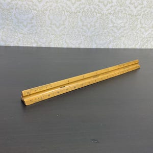 Peut inclure: Une règle d'architecte ancienne en bois. La règle est de couleur marron clair avec des marquages et des chiffres noirs. Elle comporte plusieurs échelles pour différentes mesures. La règle mesure environ 30 cm de long.