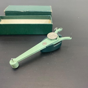 Könnte beinhalten: Ein grünes und silbernes Präzisionsinstrument aus der Zeit, möglicherweise zum Messen oder Markieren, mit einem Zifferblatt und zwei scharfen Spitzen. Das Instrument wird von einer grünen Box mit Deckel begleitet. Das Werkzeug wird wahrscheinlich für technische oder wissenschaftliche Zwecke verwendet.