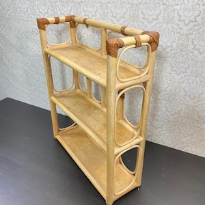 Peut inclure: Une étagère à trois niveaux en rotin brun clair, dotée d'un cadre rectangulaire aux coins arrondis et de liens tressés décoratifs aux jointures. La structure se compose de trois tablettes plates soutenues par des montants verticaux.