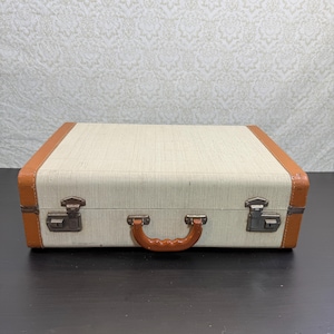 May include: A rectangular suitcase featuring a textured off-white centre panel and tan-coloured edges. The suitcase includes a central tan handle, two metal latches with keyholes on the front, and metal hinges along the bottom edge.