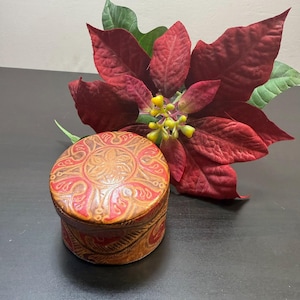 Peut inclure: Une boîte ronde en cuir avec un motif floral sculpté sur le couvercle et les côtés. La boîte est principalement marron avec des accents rouges. Une grande fleur de poinsettia rouge avec des feuilles vertes est en arrière-plan.
