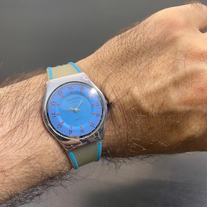 Puede incluir: Un reloj de pulsera Swatch con caja plateada y esfera azul claro que muestra los números del 13 al 24 en disposición circular. El reloj tiene una correa de silicona beige con bordes azul brillante y el texto Swatch Swiss Quartz en la esfera.