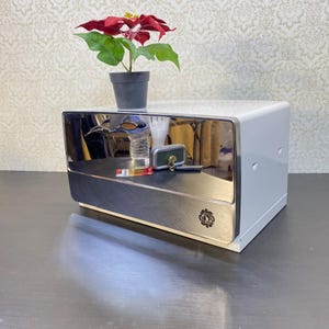 May include: A white and silver metal box with a reflective front panel. A small potted plant with red and green leaves sits on top. Keys and other small items are on the box's surface.