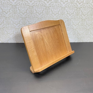 May include: A light brown wooden cookbook stand. The stand has a sloped design with a ledge to hold a book and a curved top. The stand is made of wood and is designed to hold cookbooks or other reading materials. The stand is approximately 25 cm in height.
